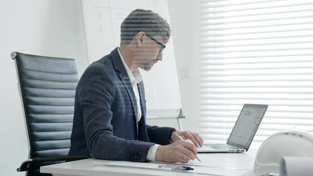 Architect or engineer working on construction blueprints and plans at an office desk, using a laptop for design and planning, embodying professionalism and project management