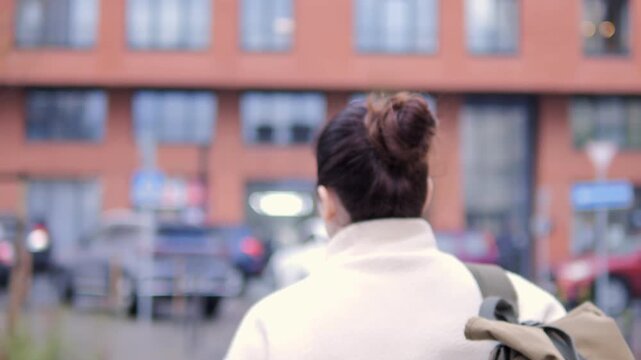 Woman with backpack walks away from camera down urban street. Hair in bun, wearing light-colored coat. Blurred cars and brick building in background. Calm. Suitable for scenes of urban life