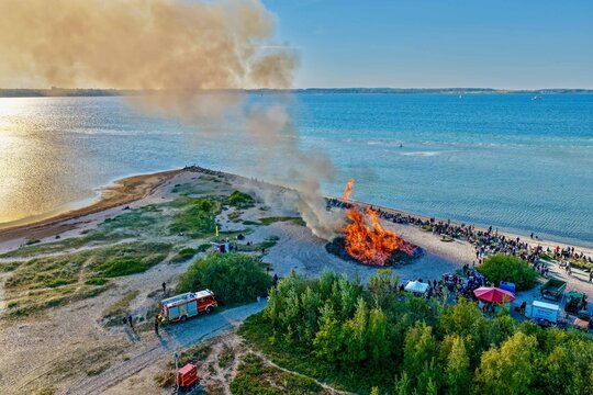 Laboe, Germany - 09 May 2026: Aerial view of a large Maifeuer bonfire burning on the beach with a crowd of people and a fire truck nearby.