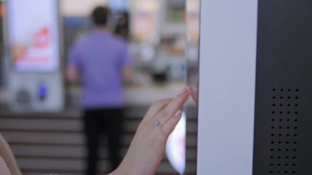 Person interacts with a modern touchscreen kiosk. Their hand gently taps the screen, focused and deliberate. Background shows blurred indoor space with other people. Footage ideal for retail
