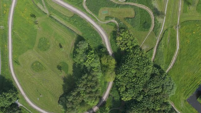 Top down aerial drone footage flying above a green park landscape with winding paths, trees, grass and small water features. Peaceful outdoor scene with natural patterns, curves and recreational infra