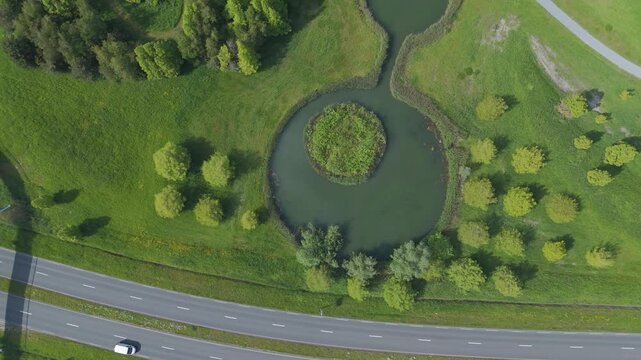 Top down aerial drone footage flying above a green recreational park landscape with ponds, trees, grass, winding paths and a road with passing cars. Peaceful outdoor nature area with geometric paths.
