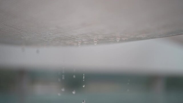 Cinematic horizontal close up view of water dripping through a canvas awning or fabric shade sail in heavy rain with soft blurred background - 비 내리는 날 야외 테라스의 천막 어닝이나 그늘막 천 사이로 맑은 빗방울이 송글송글 맺혀 아래로 톡톡 