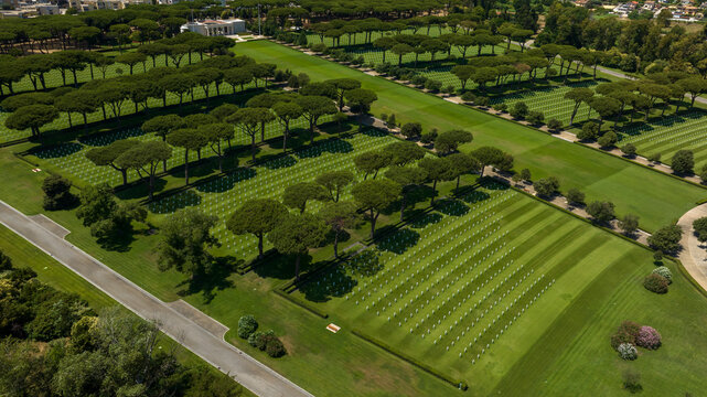 Aerial view of a military cemetery, featuring meticulously manicured green lawns with distinct striped patterns, lined by parallel rows of tall umbrella pine trees. It's located in Nettuno, Italy.