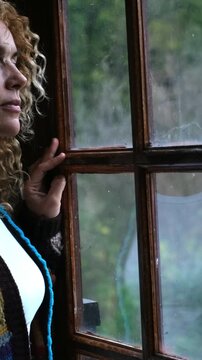 Lonely adult caucasian woman inside cabin chalet home looking outdoor through the glass window. Concept of loneliness and sadness for female people. Stress and independent lifestyle lady