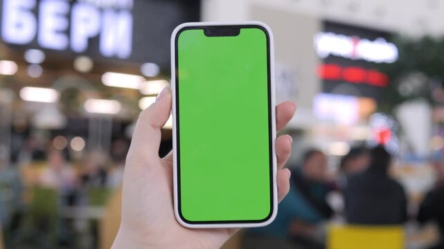 Hand holds smartphone with green screen. Background shows blurred shopping center. Focus on phone for overlay use. Ideal for ads, apps, or UI design projects