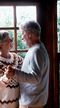 Happy senior couple in love dance at home agains a big windows view with nature woods outside. Romance leisure activity mature man and woman elderly retired lifestyle