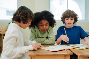 Fototapeta premium Diverse students sitting at classroom desk reading book and writing with pencils during school lesson within educational environment