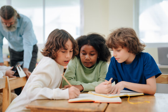 Diverse students sitting at classroom desk reading book and writing with pencils during school lesson within educational environment