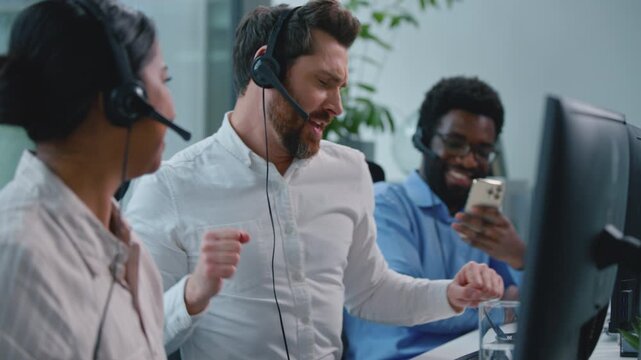 Three multiethnic call center operators business team. Caucasian agent crazy happy singing dancing fun at table in callcenter African American man recording video with mobile phone Arabian woman dance