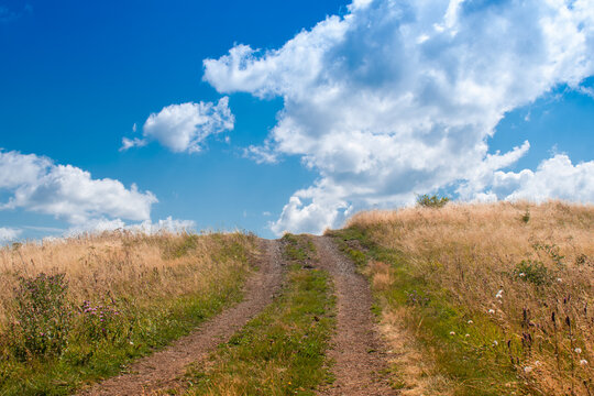 Dirt road leading up a grassy hill under a beautiful blue sky with fluffy white clouds. Countryside dirt track through a golden field on a sunny summer day. Rural road to the sky.