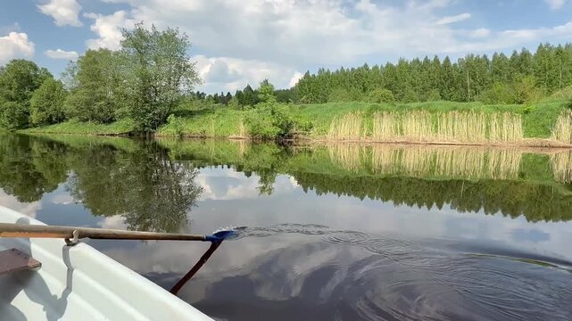 A view from a rowboat shows an oar dipping into a calm river, reflecting green trees and a cloudy sky.
Concept of peaceful summer boating and nature exploration.