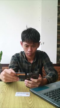 Young man sitting uses a credit card and smartphone with a laptop nearby in a casual setting