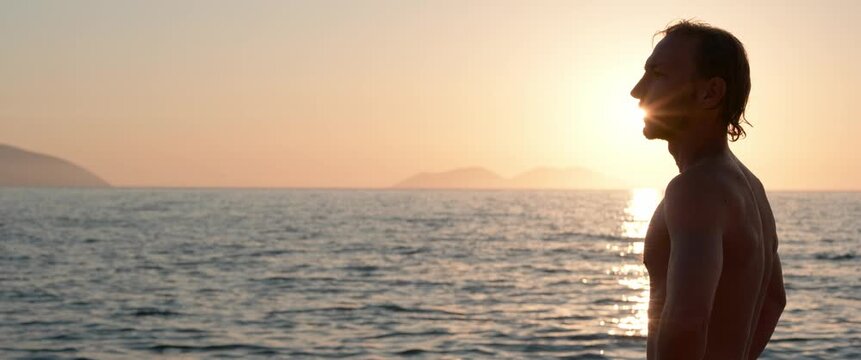 Man silhouetted at the edge of the frame gazing at the sea and distant hills during sunset, anamorphic static wide shot.