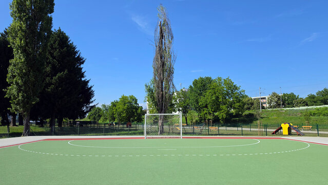 White metal soccer goal with net on a green hardcourt sports field bordered by trees under a blue sky