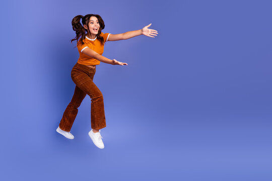 Happy young girl jumps in orange top and brown pants against violet background for lifestyle fashion promotion