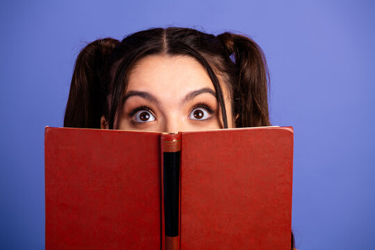 Young female teen girl peeking over a red book with a curious smile for study and fun lifestyle