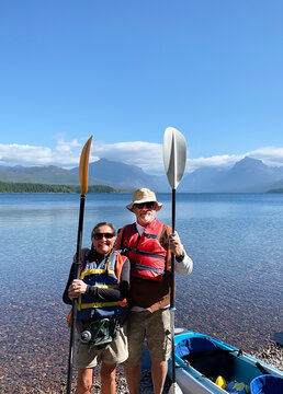 Smiling couple  beside their kayak on Lake McDonald in Glacier National Park, Wyoming, USA enjoying an outdoor adventure, summer travel and peaceful nature scenery with distant peaks and calm water.