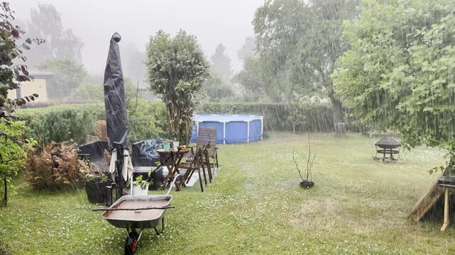 A heavy rainstorm sweeps across a backyard with a blue pool, patio table, umbrella, and wheelbarrow. Wet grass, hedges, and garden trees show intense summer weather.