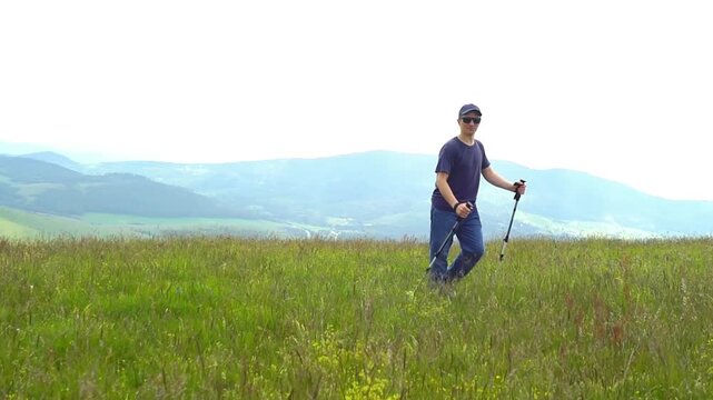 Laughing hiker with trekking poles turns from camera and starts walking across windswept mountain meadow. Portrays enjoyable outdoor recreation, healthy leisure habits, and appeal of highland hiking.