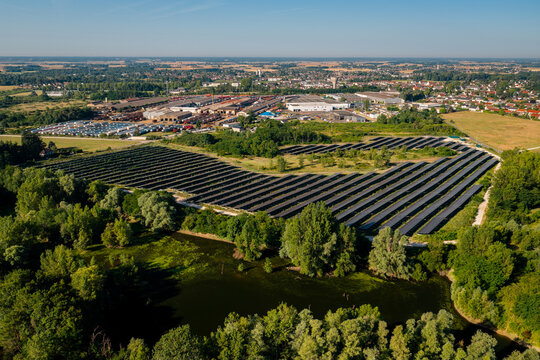 Aerial view of a solar farm with rows of photovoltaic panels near a pond and trees, with an industrial area and the town of Saint-Denis-de-l'Hotel, Centre-Val de Loire, France.