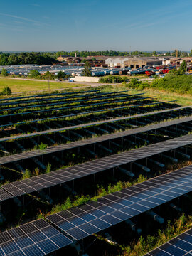 Aerial view of solar panels arranged in rows over a grassy field with a rail yard and industrial buildings in the background in Saint-Denis-de-l'Hotel, Centre-Val de Loire, France.