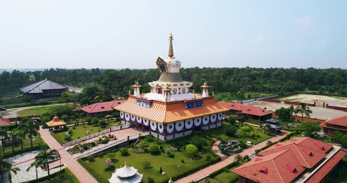 Hazy aerial drone view of a golden roofed Buddhist temple decorated with white circular motifs, set among palm trees and green gardens in Lumbini.
