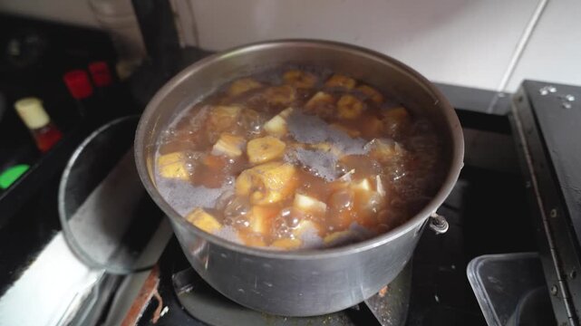 Boiling Indonesian sweet dessert kolak with sliced bananas and cassava in a pot on a stove, close up high angle view.