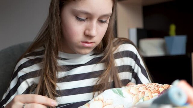 Teenage girl sitting at table and sewing by hand with needle and thread. Focused young seamstress doing handmade needlework at home.