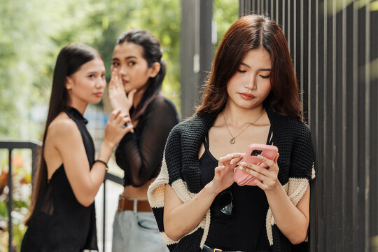 Two girls whisper and gossip behind a woman focused on her phone, capturing social judgment, exclusion, and subtle tension.