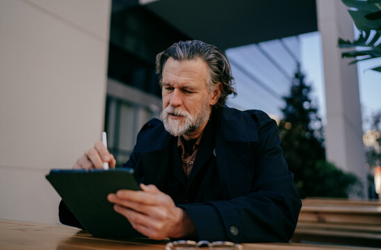 Creative senior man sketching on tablet with stylus, mature professional developing application or writing notes at outdoor workspace, digital nomad, online learning, it professional, technology.