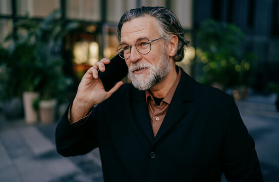 Smiling mature gentleman having pleasant mobile call outdoors, senior man in glasses and dark coat phoning in city, positive attitude, 5G wireless communication, authentic representation, lifestyle.