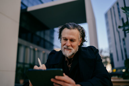 Focused senior designer drawing on digital tablet with stylus, mature creative professional using touch screen for project planning at outdoor cafe, technology, startup culture, real people.