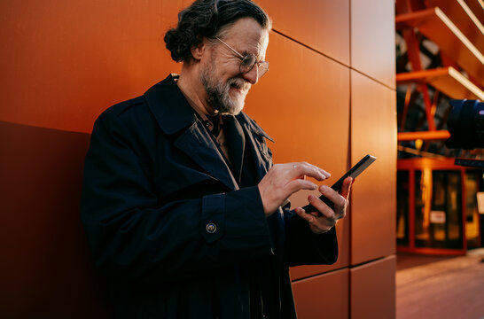 Smiling senior man using touchscreen of smartphone for booking or online search, mature businessman in black coat holding mobile phone against modern office wall during sunset.