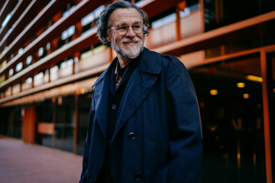Portrait of cheerful senior businessman in black trench coat standing outdoors, happy mature male manager posing in front of modern glass office building in urban area.