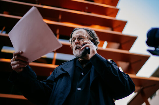 Angry mature businessman holding documents and talking on smartphone outdoors, senior professional male manager having emotional mobile conversation in urban area.