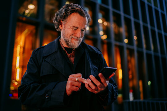 Successful mature male professional using smartphone for business tasks, senior man with beard typing on mobile phone inside contemporary office lobby, digital nomad lifestyle with copy space.