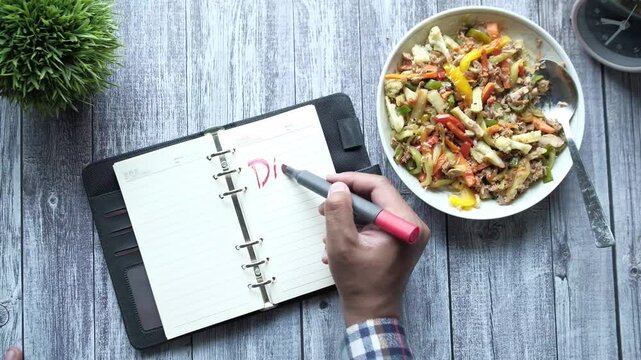 Writing diet plan in notebook with healthy food beside on wooden table