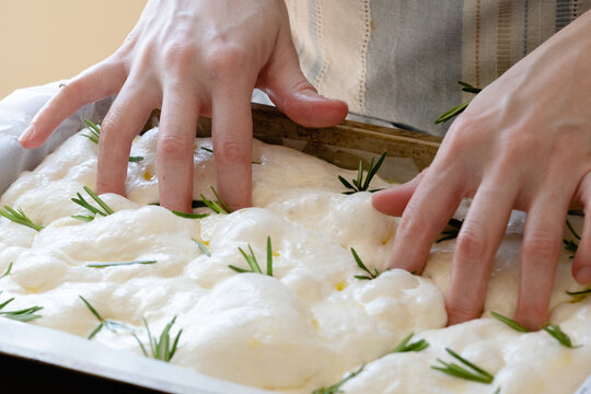 Unrecognizable female's hands gently pressing down on soft, risen dough in a baking tray. Fresh rosemary is placed on top as the baker prepares to create focaccia. Traditional bread making