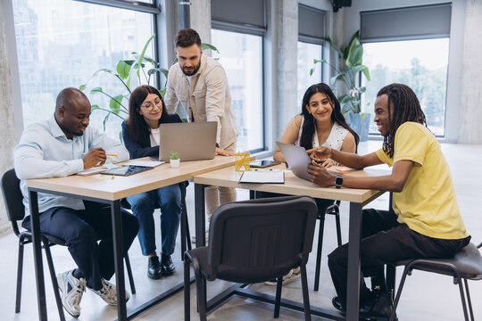 Diverse business people gathering around tables, interacting and using laptops in a bright, modern office environment