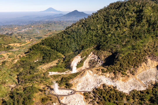 Aerial view of volcanic steam vents rising from a lush green forested mountainside with distant peaks in the background in Indonesia.