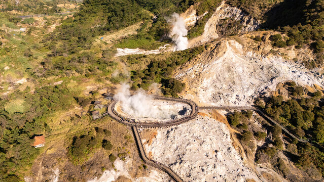 Aerial view of the steaming geothermal crater of Kawah Sikidang with a wooden boardwalk winding through the rocky volcanic landscape surrounded by lush green hills in Indonesia.