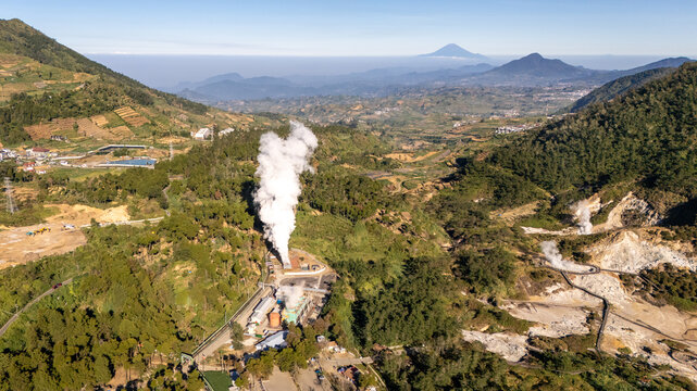 Aerial view of a geothermal power plant emitting a large white steam plume amidst lush green mountainous terrain and volcanic landscape in Indonesia.