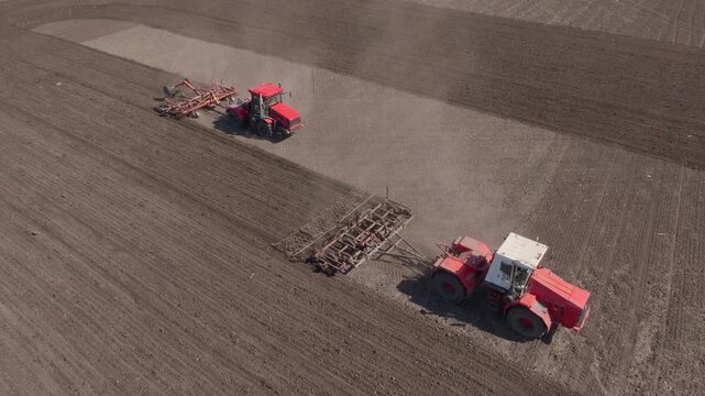 Aerial drone view of two red tractors pulling wide harrows across a dry field, creating parallel cultivation passes and raising dust clouds during tillage on expansive farmland