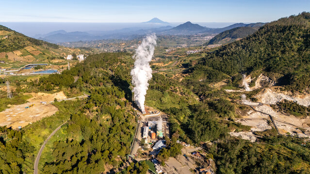 Aerial view of a geothermal power plant with a prominent steam plume rising above lush green mountainous terrain in Indonesia.