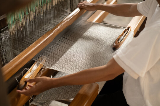 Over the shoulder view of a skilled artisan operating a wooden handloom, weaving beautiful grey wavy patterned tenun fabric. Authentic Indonesian textile art.