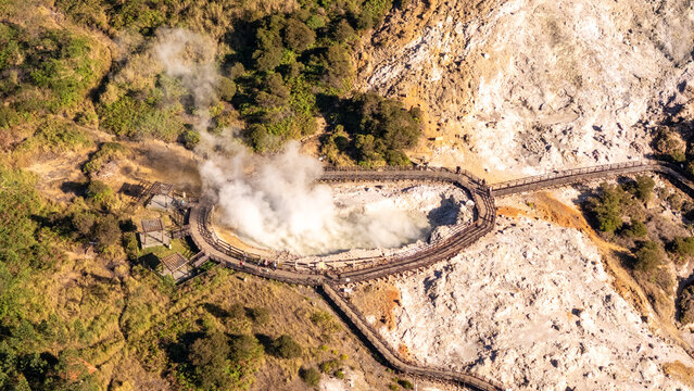 Aerial view of the steaming Sikidang Crater with its wooden boardwalk winding through the rocky geothermal landscape in the Dieng Plateau, Indonesia.