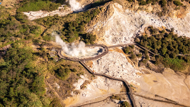 Aerial view of a steaming volcanic crater surrounded by lush green forest and wooden boardwalks in Indonesia.