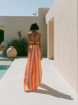 Woman in a striped maxi dress walks away from the camera beside a swimming pool, with a modern architectural backdrop and greenery in the scene