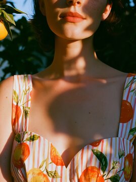 Woman with short hair wearing a citrus-patterned dress stands in front of a lemon tree, sunlight casting shadows on her shoulders and neckline